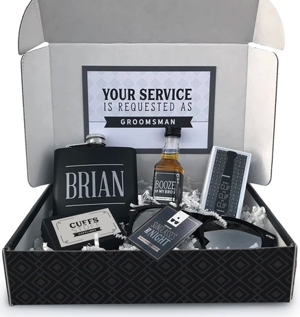 Black box with a personalized flask, whiskey, cufflinks, sunglasses, and a tie clip, asking "Will you be my groomsman?"